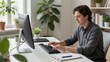 © BOJOShop - Young man working at desk with desktop computer and keyboard in bright modern home office with green plants and stationery for remote work and digital productivity concept