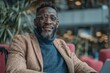© Alexei - African man wearing glasses and a brown blazer smiles while seated in a modern indoor setting with greenery and soft seating visible in the background