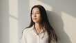© Stunning stock - Serene Asian woman in white linen shirt leaning against wall, thoughtful young woman in natural sunlight, minimalist portrait with soft shadows