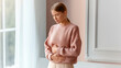 © dinatychynska - Young woman holding abdomen near window, menstrual cramps, stomach pain concept. Digestive discomfort, period pain, gut health, calm pastel interior, lifestyle.