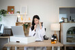 © Stella 16 - Young Asian woman doctor in white coat multitasks on phone call while writing on clipboard at Medical Office Desk, ideal for healthcare and telemedicine concepts