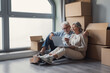 © Daniel - Senior couple sitting on the floor of their new apartment among cardboard boxes, smiling and using a laptop to plan moving, unpacking and home arrangements together