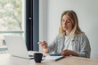 © Daniel - Young woman working productively at a desk, looking at her laptop screen and holding a pen while learning or studying in a brightly lit home environment