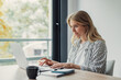 © Daniel - Young woman working from home on a laptop at a desk by a window, notebook and coffee nearby, focused on remote freelance tasks in a bright, casual home office setting