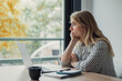 © Daniel - Young adult woman with blonde hair wearing a striped shirt sitting at a table with a laptop and coffee cup, looking out the window, appearing bored or lost in thought