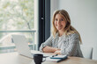 © Daniel - Young woman smiling confidently at her desk with laptop, notebook, pen and coffee, embodying productive remote work lifestyle, modern freelance professional in home office