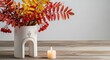 © suchetpong - A minimal white ceramic vase filled with colorful fall branches and red berries stands on a wooden table beside a glowing candle flame, aesthetic, warm, soft