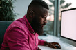 © Shelley - Focused Black Man in Pink Shirt Working at Desk with Large Monitor in Bright Office with Natural Light and Plants.