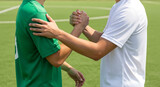 Two soccer players wearing green and white jerseys shaking hands on a green grass field, showing sportsmanship and respect after a competitive match