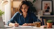 © Pcs - A young woman wearing a blue shirt, sitting at a desk with a laptop and a cup of tea, writing in a notebook.