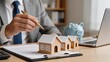 © Aiden - Real estate investment planning with miniature houses on clipboard, businessman in suit holding gold pen, piggy bank and laptop in background, property management or mortgage