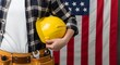 © Creatives - Labor Day Construction worker holding a yellow hard hat with a tool belt against a blurred American flag background