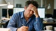 © ASyen - A stressed businessman holding his head while sitting at a desk in an office with a computer in the background, looking unwell