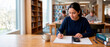 © Xenia - Female university student studying math in the library. Young woman writing in a notebook and using a calculator at a desk. Higher education concept
