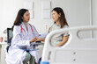 © Wasan - Young Asian female doctor in white coat with stethoscope smiling and explaining medical information to older Asian female patient seated on hospital bed during clinic visit.