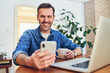 © baranq - Man smiling using smartphone and laptop at home