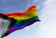 © rarrarorro - Progress Pride Flag ripples in the wind against a blue sky with soft clouds. 3D rendered illustration of the inclusive LGBTQ+ symbol for diversity and equality.