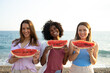 © SISMIZ - Three smiling women holding watermelon slices at the beach during a sunny day