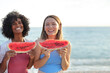 © SISMIZ - Two smiling women enjoying watermelon slices at the beach on a sunny day