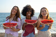 © SISMIZ - Three diverse women laughing and holding watermelon slices at the beach