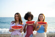 © SISMIZ - Three smiling friends holding watermelon slices at the beach on a sunny day