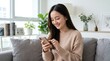 © MF - 明るくモダンなリビングルームでソファに座ってスマートフォンを使う幸せなアジアの若い女性