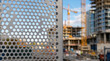© ViskBx - Perforated metal sheet fence in front of a construction site with cranes and unfinished highrise buildings in the background