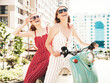 © halayalex - Two young beautiful smiling hipster female in trendy overalls.Sexy carefree women driving retro motorbike on the street background.Positive models having fun, riding classic Italian scooter in eyewear