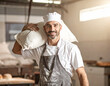 © magele-picture - Ein Bäcker trägt einen großen Sack Mehl in der Bäckerei