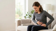 © Xenia - Woman sitting on sofa holding stomach in pain. Female suffering from abdominal discomfort or menstrual cramps at home. Healthcare and medical concept