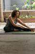 © wavebreak3 - African American woman sitting on green yoga mat at patio stretching in sportswear with smartwatch