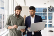 © fizkes - Two multinational male colleagues engaged in discussion in office boardroom, working with tablet and laptop, share ideas, reviewing or comparing data, take part in efficient teamwork, digital workflow
