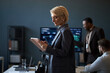 © DragonImages - Middle aged Caucasian businesswoman reviewing tablet during team briefing in office, managing digital workflow and leadership strategy. Ideal for corporate communication and management