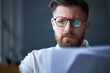 © DragonImages - Close up of Caucasian young adult businessman reviewing documents through eyeglasses, focusing on office task and decision making. Suitable for business planning or finance content