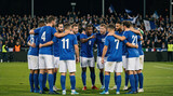 Professional male soccer team gathering in a huddle on the field. Football players in blue and white uniforms discussing game strategy under stadium lights. Teamwork and athletic competition concept