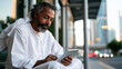 © Kateryna - A businessman in a soft crisp thobe waits softly at a calm modern bus stop reading a bright tablet in soft daylight bus stop wait modern transit thobe attire tablet read