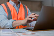 © Teelapon - Close-up of an industrial worker wearing high visibility safety vest using laptop computer and reviewing documents in office or construction workplace environment.