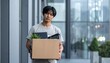 © photostockatinat - A sad male employee wearing a casual t-shirt holding a cardboard box of personal belongings after a sudden company layoff notice.