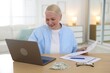 © New Africa - Pension. Senior woman with paperwork, money and laptop at table indoors