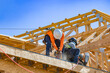 © Andrei Liashchou - A wide horizontal view showing two construction workers wearing safety harnesses and hard hats while installing and securing wooden roof trusses on a new building framing structure under a clear blue