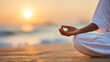© Oksana - A person in white clothing sits in a meditation pose on a beach at sunset, with a focus on their hand in a mudra gesture against the softly blurred ocean horizon.