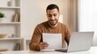 © 5 Grace Media - Smiling young man reading document at home office desk with laptop.