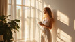 © Martarina - Serene young woman holding a cup of coffee or tea standing by the window with soft morning sunlight and shadows