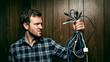 © Shyla Marsare - A stressed adult man struggles with a chaotic tangle of various electronic cables, USB cables, and chargers in a dark room. Conceptual image of technological frustration and computer problems.
