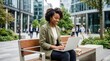 © ZHMPRO - Professional african american woman working on laptop computer while sitting on outdoor park bench in modern corporate city area.