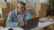 © Krakenimages.com - Middle aged man with grey hair working on laptop, hand on chin, surrounded by packed cardboard boxes in building; concern moving transition.