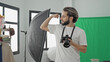 © Krakenimages.com - Man photographer holding camera flexes bicep in a studio with green screen and lighting equipment; creative confidence.