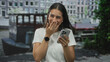 © Krakenimages.com - Young brunette woman holding smartphone, hand to mouth laughing while checking messages and swiping feeds on street by canal boats and a riverside building; joy.