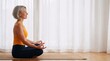 © Przemek - Peaceful mature woman with short grey hair practicing meditation and mindfulness on a yoga mat in a bright, sunlit room with white curtains.