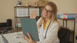 © Krakenimages.com - Woman doctor waving at tablet in medical office building, wearing white coat and stethoscope, smiling during virtual consult; telehealth connection reassurance.